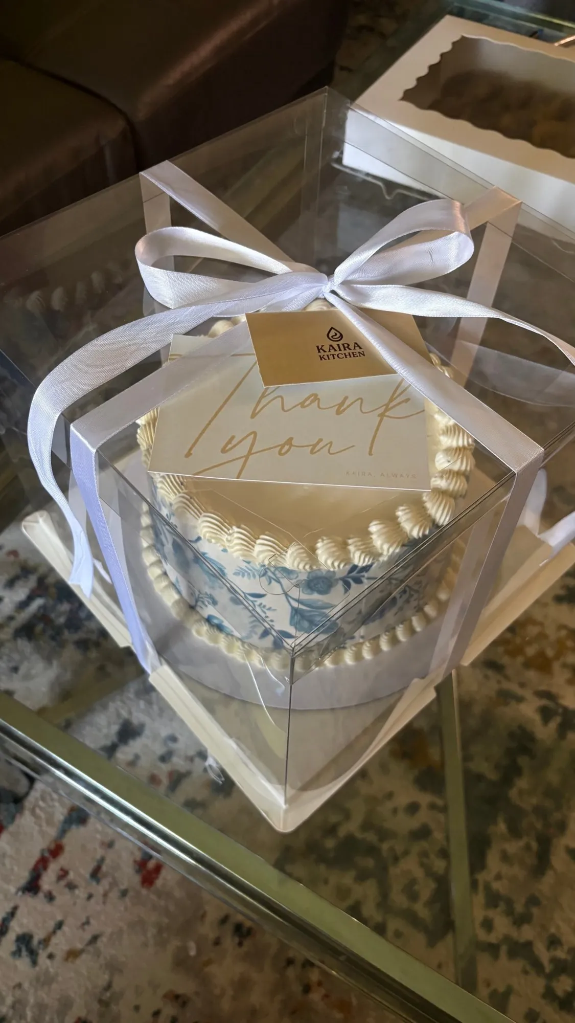 Cream buttercream cake with blue floral pattern, packaged in a clear gift box with ribbon and a Kaira Kitchen thank-you card