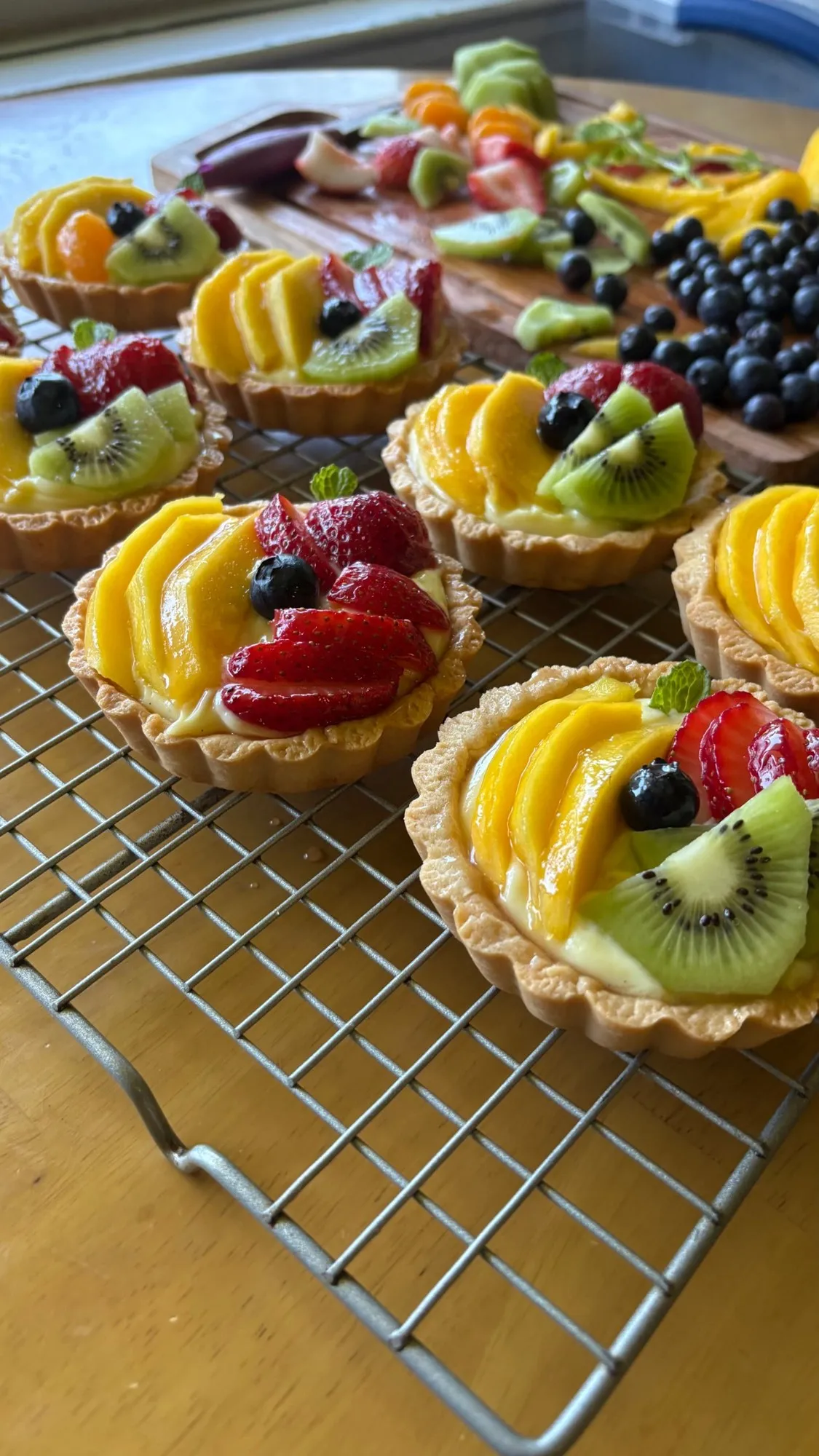 Mini fruit tarts on a cooling rack with fresh mango, strawberries, kiwi, and blueberries