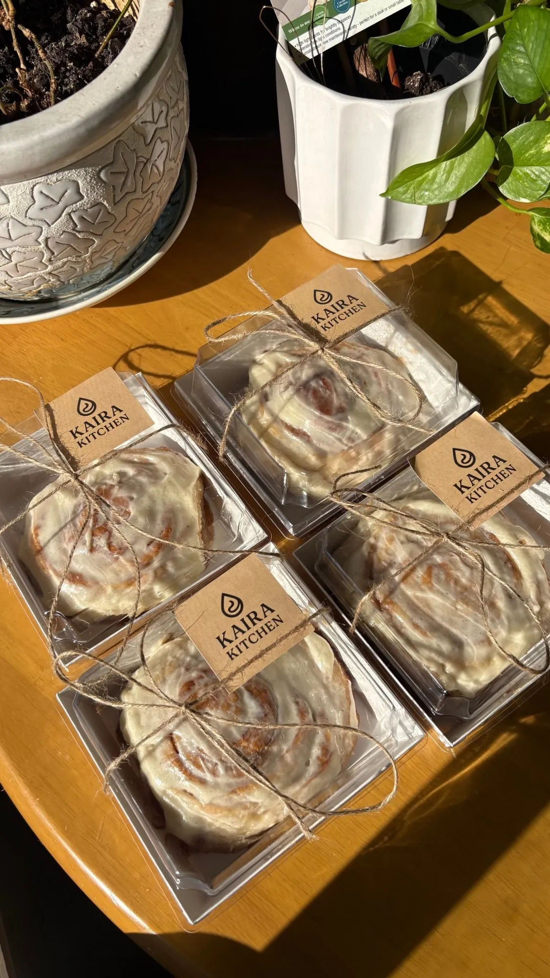Four iced cinnamon rolls in clear boxes tied with twine and Kaira Kitchen kraft labels